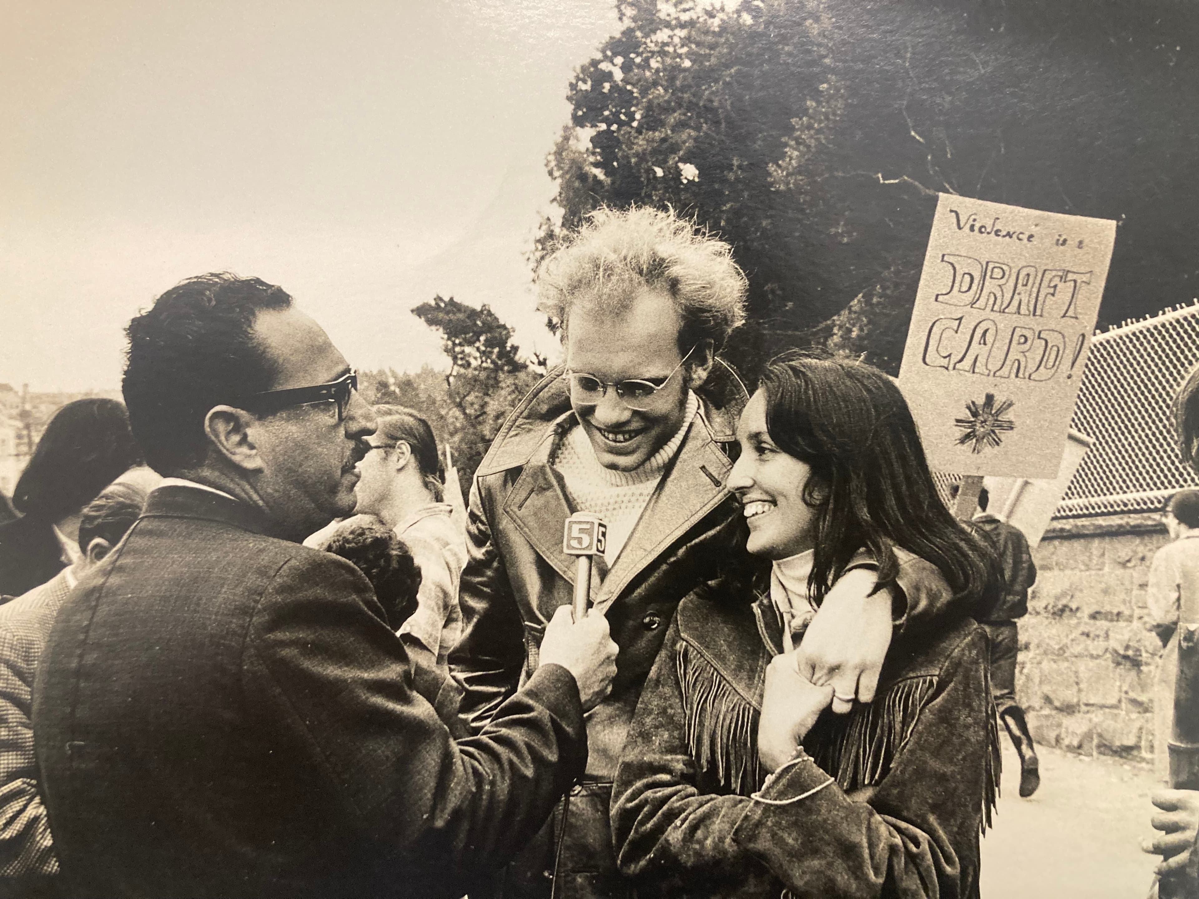 A sepia photograph of David Harris with his arm around Joan Baez, both smiling while speaking to a reporter at an anti-war protest rally.