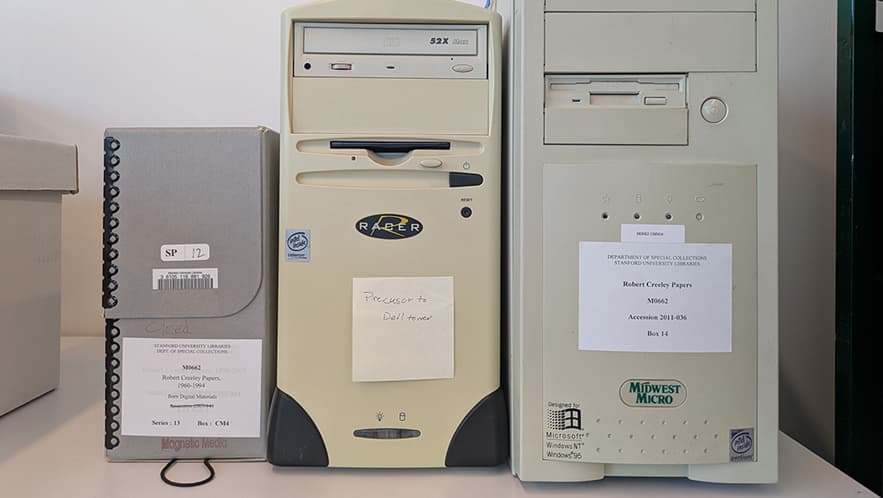 One light gray archival Hollinger box and two off-white desktop computer towers (one Racer desktop PC and one Midwest Micro PC) are lined up on a table.