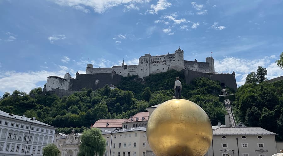 A large golden orb with a male figure on top, situated in a town square with a mountaintop fortress in the background.